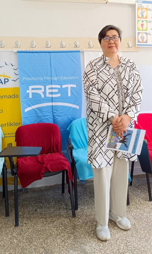 A middle-aged woman with glasses stands confidently in front of a RET banner, wearing a patterned coat and holding a booklet, with colorful chairs in the background.