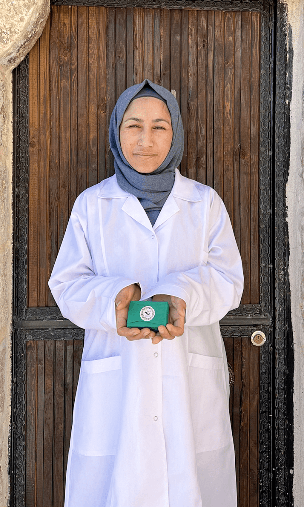 A woman wearing a white lab coat holds a green box with a logo in front of a wooden door.