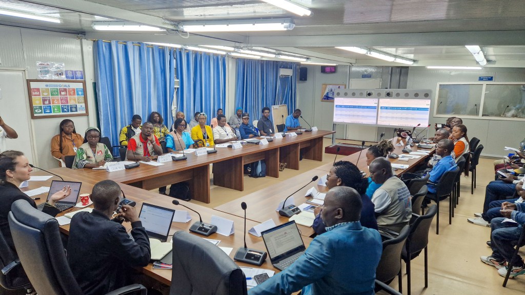 A group of diverse individuals participating in a meeting around a long wooden table in a conference room, with blue curtains and screens displaying information in the background.