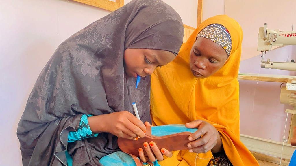 Two women, one in a black hijab and the other in a yellow hijab, collaborate on a shoe-making project. The woman in black is drawing on a shoe sole while the woman in yellow observes attentively. A sewing machine is visible in the background.