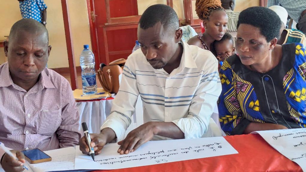 Three people seated at a table engaged in writing on papers during a workshop. Two men and a woman are focused on their tasks, with a water bottle and a phone visible on the table.