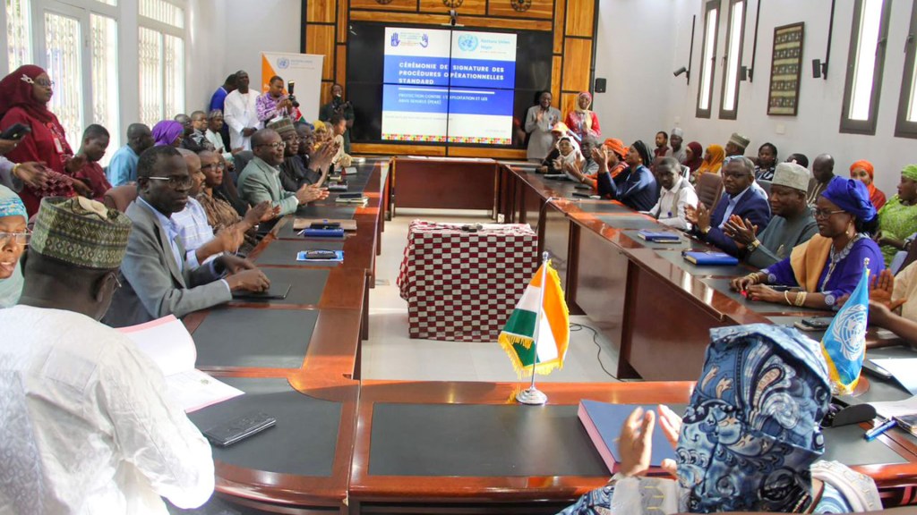 A formal ceremony scene with attendees seated around a large table, some clapping, in a conference room. A screen in the background displays information about the event, and flags are visible on the table.
