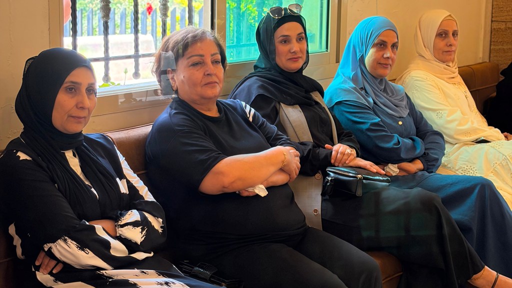 Five women sitting side by side on a couch, dressed in various styles of traditional and modern attire, with a window in the background.