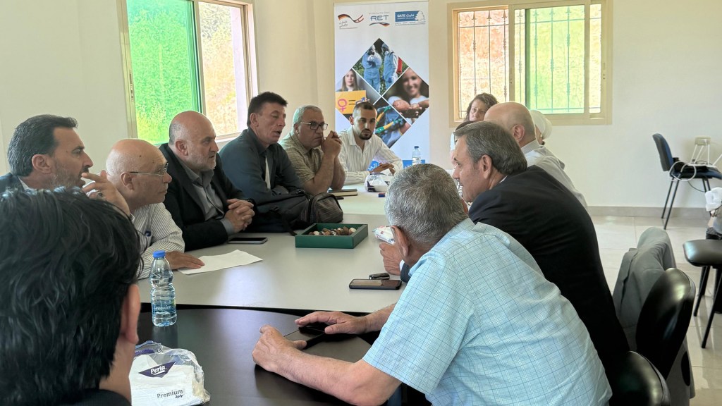 A group of men engaged in discussion around a conference table in a well-lit room. Several participants are leaning forward, actively communicating. In the background, there is a promotional banner with images and text.
