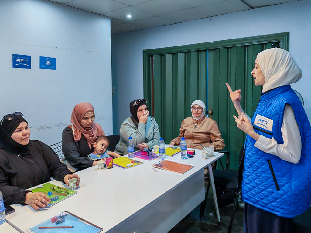 A group of women seated around a table in a classroom setting, listening attentively to a female speaker wearing a blue vest. The room features a green door and several bottles of water on the table.