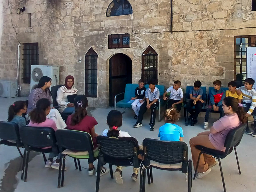 A group of children and adults seated in a circle in front of a rustic stone building, engaged in a discussion or activity.