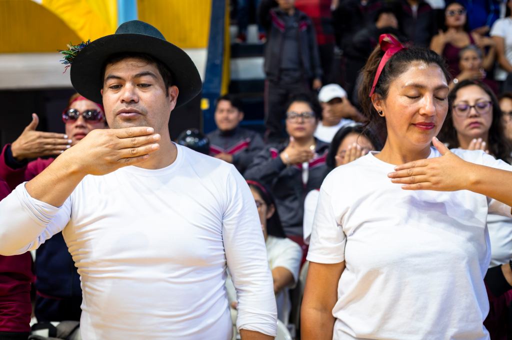 Participants performing a synchronized gesture during an event, with one man wearing a hat and a woman in a headband, both in white shirts, surrounded by an audience in a gymnasium.