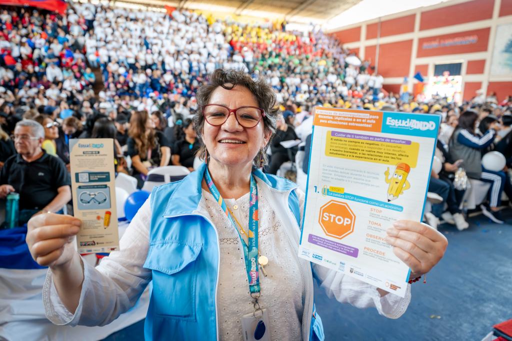 A smiling woman wearing a blue vest holds two informational flyers in a crowded indoor venue. The background is filled with people in various colored shirts, some holding balloons.