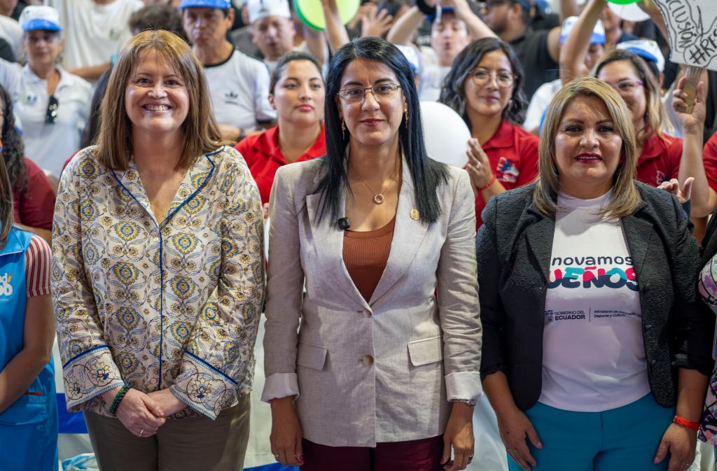 Three women stand together in a crowded event, smiling for the camera. They are dressed in formal attire, with one in a patterned blouse, another in a light-colored suit, and the third in a black jacket with a white shirt. The background shows a group of attendees, some holding up signs and balloons.