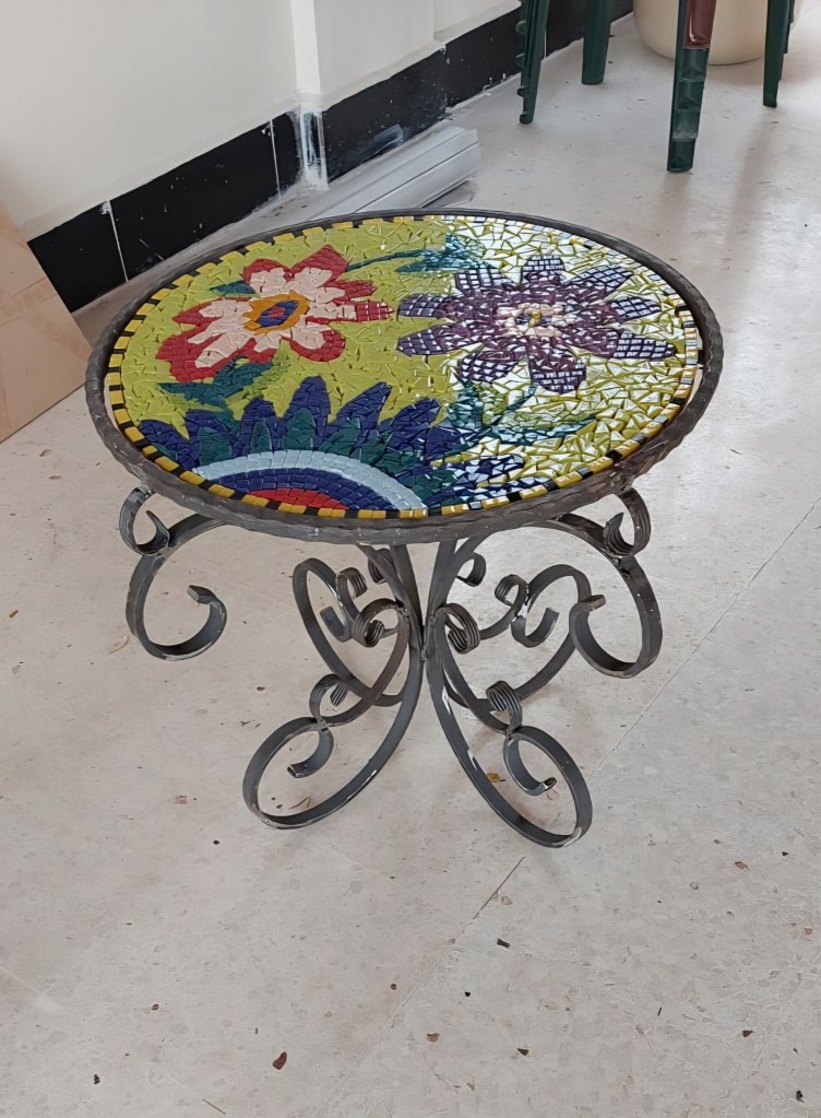 A round table with a colorful mosaic top featuring floral designs, supported by an ornate black metal base.