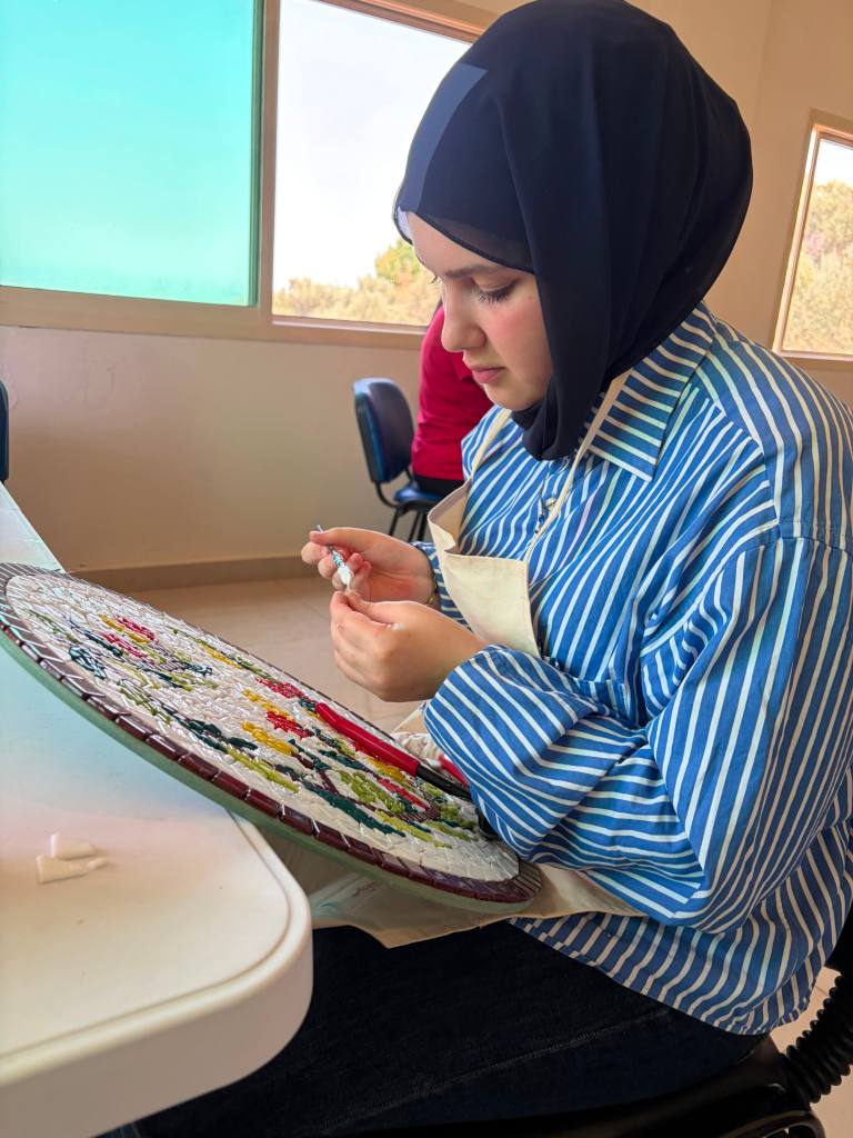 A young woman wearing a hijab and a striped shirt is focused on crafting a mosaic art piece, holding a small tile in her hand while seated at a table in a well-lit room.