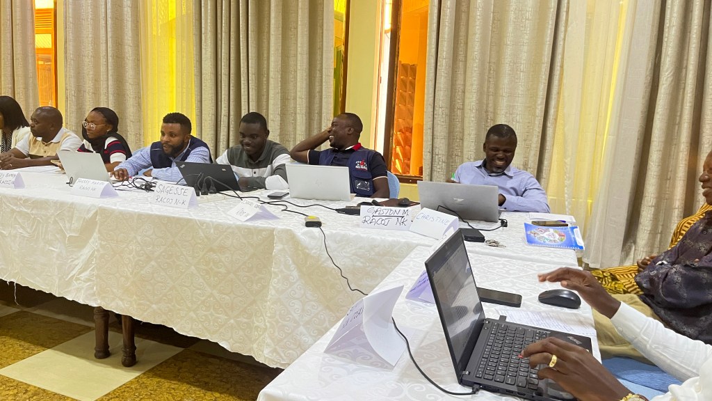 A group of people sitting around a table during a meeting, each with laptops. The scene includes a selection of attendees engaging in discussion, with some people showing smiles and others focusing on their computers.