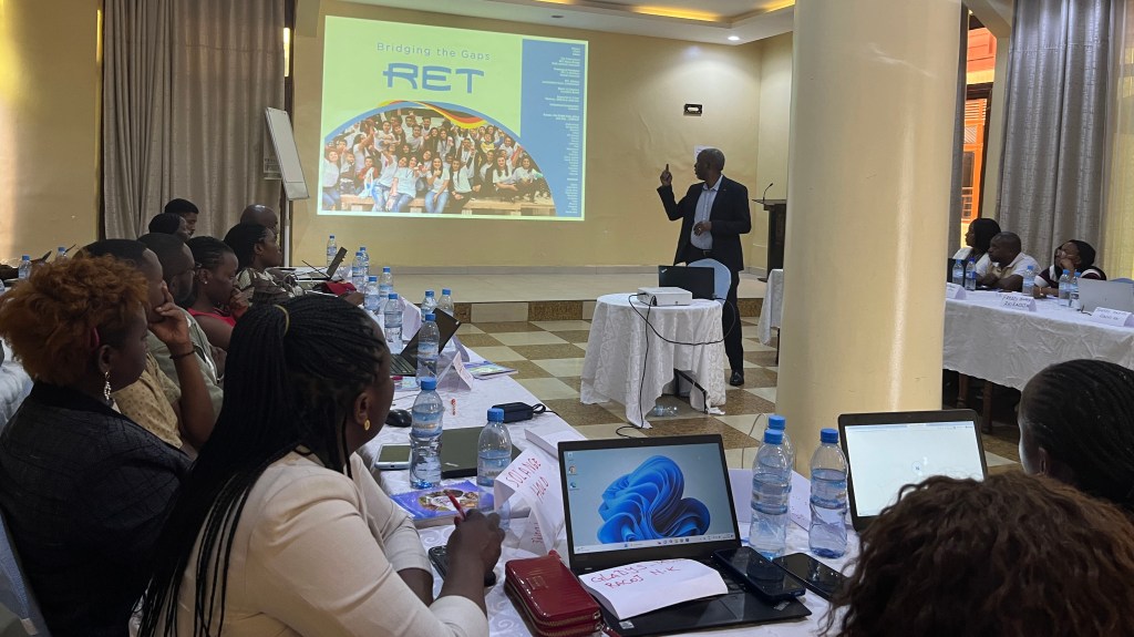 A speaker presenting in front of an audience at a conference, with a slide titled 'Bridging the Gaps' and a group photo on display. Attendees are seated at tables equipped with laptops and water bottles.
