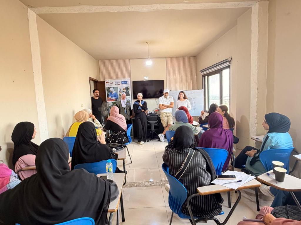 A group of women seated in a classroom setting, engaging with two presenters standing at the front. There are various notes and materials on the desks, and a television screen displaying information in the background.