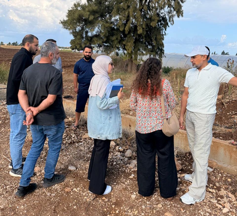 A group of six people gathered outdoors, discussing agricultural practices near a farm. They're standing by a tree, with a blue sky and clouds in the background.