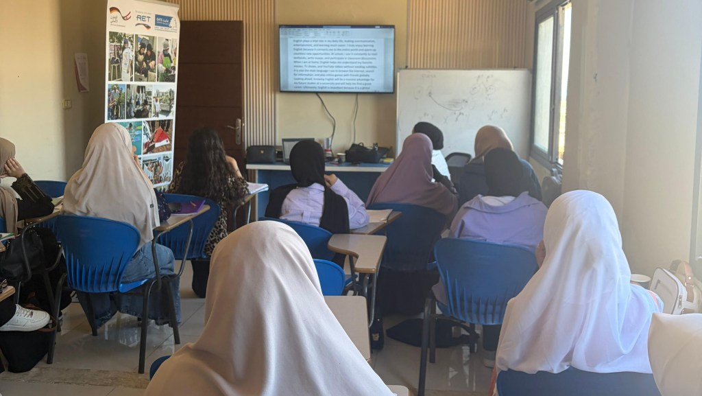 A classroom setting with students wearing hijabs seated at desks, attentively watching a presentation displayed on a screen at the front of the room. A banner showcasing various activities is visible on the left side.
